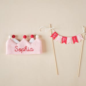 K&ouml;nnte beinhalten: Rosa Filzkrone mit roten Filzkugeln und dem Namen "Sophia" in roter Farbe gestickt. Ein rosa und rotes Filzbanner mit dem Namen "Sophia" auf einem Holzstab.