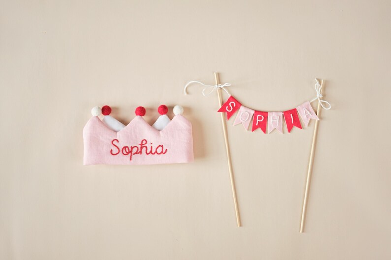 K&ouml;nnte beinhalten: Rosa Filzkrone mit roten Filzkugeln und dem Namen "Sophia" in roter Farbe gestickt. Ein rosa und rotes Filzbanner mit dem Namen "Sophia" auf einem Holzstab.