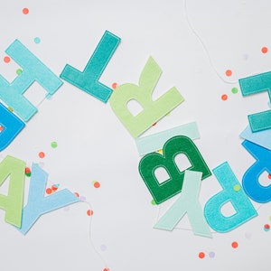 May include: A felt banner with the words "Happy Birthday" in blue, green, and light blue letters. The letters are strung together with white string and are scattered on a white background with confetti.