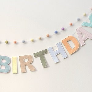 Pode incluir: Uma faixa de feltro em cores pastel que diz "BIRTHDAY" com pompons brancos enfiados entre cada letra.