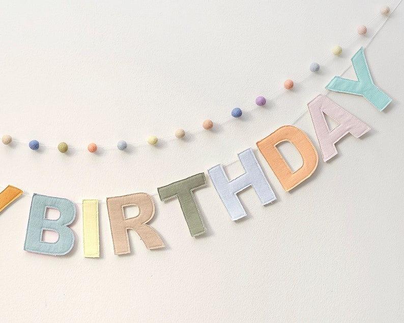 Pode incluir: Uma faixa de feltro em cores pastel que diz "BIRTHDAY" com pompons brancos enfiados entre cada letra.