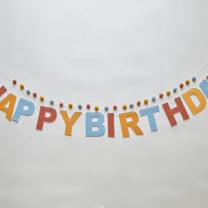 May include: A colorful felt banner that spells out "HAPPY BIRTHDAY" in orange, blue, and red letters. The banner is strung with a white cord and decorated with small, colorful pom-poms. Perfect for a birthday celebration.