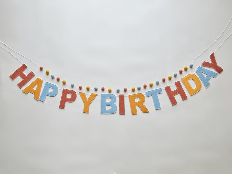 May include: A colorful felt banner that spells out "HAPPY BIRTHDAY" in orange, blue, and red letters. The banner is strung with a white cord and decorated with small, colorful pom-poms. Perfect for a birthday celebration.