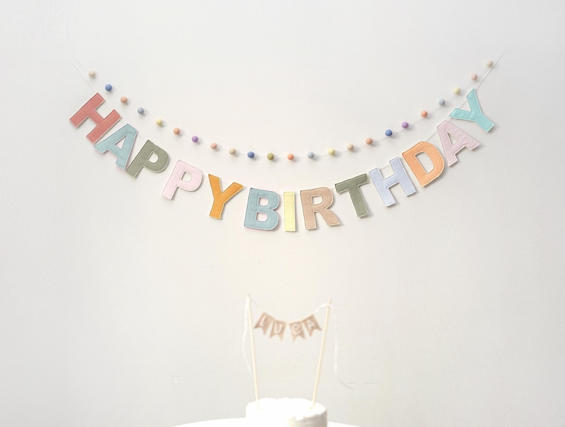 Pode incluir: Uma faixa de feltro em cores pastel com as palavras "Happy Birthday" enfiadas em uma parede branca. A faixa &eacute; decorada com pequenos pompons coloridos.