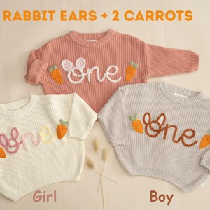 K&ouml;nnte beinhalten: Zwei Strickpullover mit gestickten Hasenohren und Karotten, einer in Rosa und einer in Grau, mit dem Wort "one" auf jedem gestickt. Der rosa Pullover ist mit "Girl" und der graue Pullover mit "Boy" beschriftet.