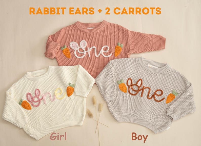 K&ouml;nnte beinhalten: Zwei Strickpullover mit gestickten Hasenohren und Karotten, einer in Rosa und einer in Grau, mit dem Wort "one" auf jedem gestickt. Der rosa Pullover ist mit "Girl" und der graue Pullover mit "Boy" beschriftet.