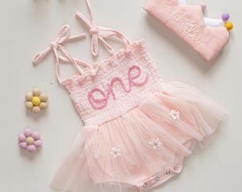 May include: A light pink baby romper with a tulle skirt and floral accents. The word "one" is embroidered on the bodice. A matching pink crown with the name "Mila" embroidered on it is also included.