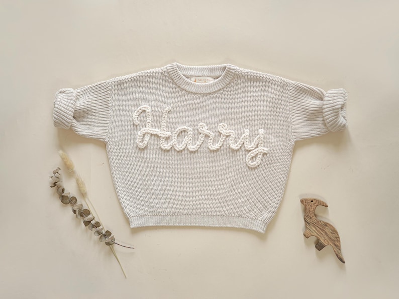 May include: A light gray knitted sweater with the name "Harry" embroidered in white yarn.