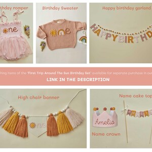 May include: A collection of birthday party decorations and apparel. Items include a pink romper with a sun design, a peach sweater with a sun design, a garland that says "Happy Birthday", a high chair banner, a crown, and a cake topper.