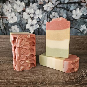 May include: Artisan soap bars in shades of pink, yellow, and green. One stack of soaps has a swirled pattern, while others are layered. The soaps are displayed on a wooden surface with a floral painting in the background.