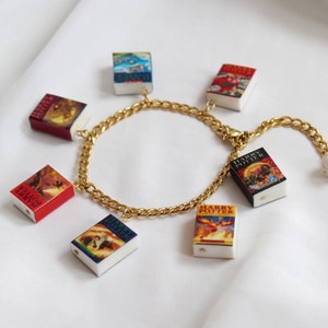 Può includere: Un braccialetto a catena dorata con ciondoli di copertine di libri di Harry Potter in miniatura. I ciondoli includono i titoli "Harry Potter e la pietra filosofale", "Harry Potter e la camera dei segreti", "Harry Potter e il prigioniero di Azkaban", "Harry Potter e il calice di fuoco", e "Harry Potter e l'Ordine della Fenice".