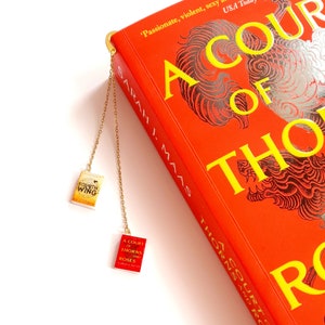 Könnte beinhalten: Eine goldene Lesezeichenkette mit zwei Miniatur-Buch-Anhängern. Die Anhänger stammen aus der Reihe "A Court of Thorns and Roses" von Sarah J. Maas. Die Anhänger zeigen die Titel "A Court of Thorns and Roses" und "A Court of Wings and Ruin".