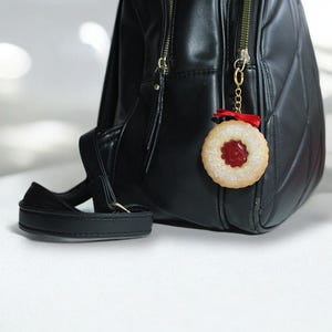 May include: A black backpack with gold zippers and a black strap. A cookie charm with a red center and a red ribbon hangs from the zipper. The charm is round with a textured edge.