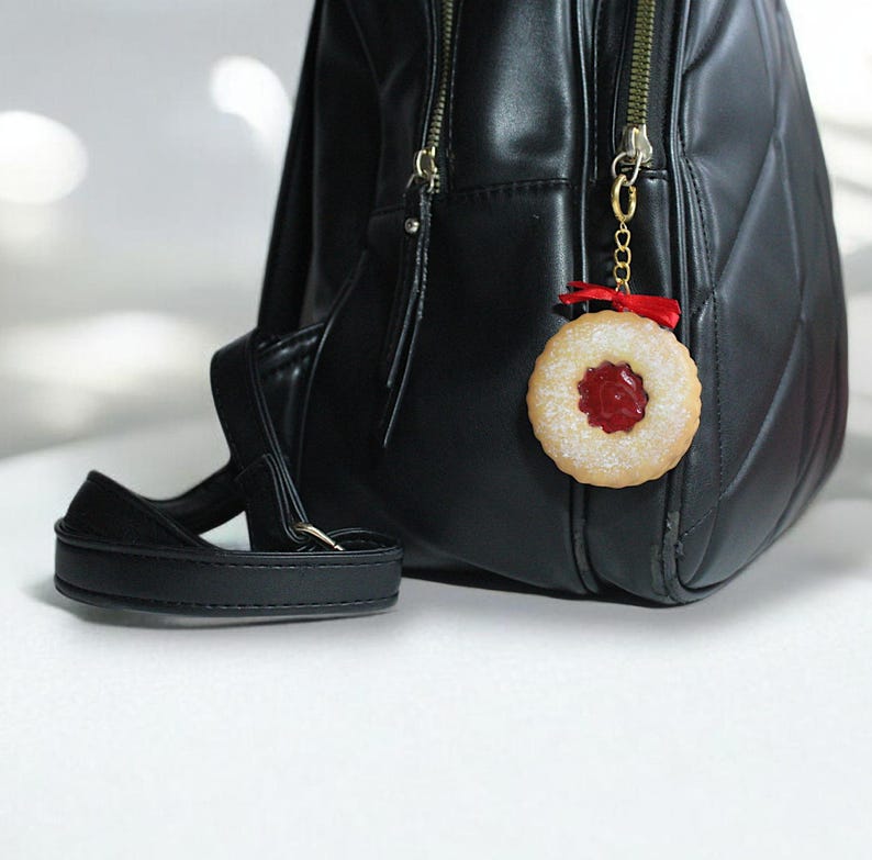May include: A black backpack with gold zippers and a black strap. A cookie charm with a red center and a red ribbon hangs from the zipper. The charm is round with a textured edge.