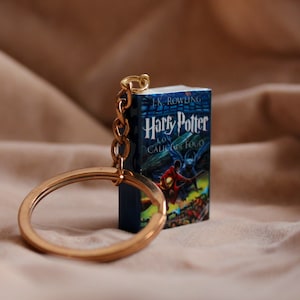 Puede incluir: Un llavero de libro en miniatura con la portada de Harry Potter y el Cáliz de Fuego de J.K. Rowling, que incluye una cadena de metal dorado y una anilla circular sujeta al borde superior del pequeño libro rectangular.