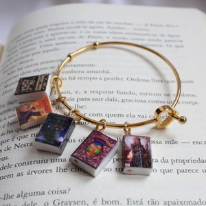 May include: A gold charm bracelet with miniature book charms. The charms feature different book covers with colorful illustrations and text. The bracelet is on a white surface with text in a foreign language.