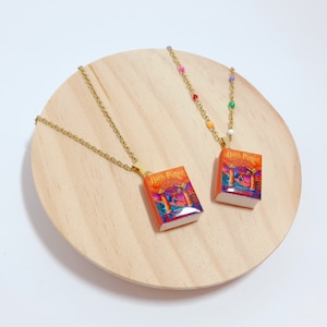 May include: Two gold necklaces with Harry Potter book charms. The charms are made of resin and feature the book cover art. The necklaces are displayed on a wooden surface.