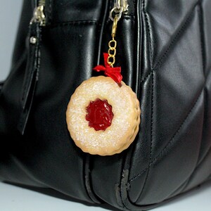 May include: A round, tan-colored charm resembling a jam-filled cookie, dusted with powdered sugar. It features a red jam center and a small red ribbon. The charm is attached to a gold-colored chain and is hanging from a black purse.