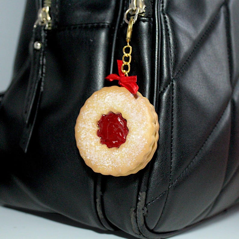 May include: A round, tan-colored charm resembling a jam-filled cookie, dusted with powdered sugar. It features a red jam center and a small red ribbon. The charm is attached to a gold-colored chain and is hanging from a black purse.