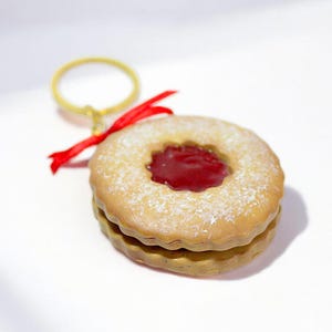 May include: A round, two-layered cookie keychain with a red jam center and a dusting of powdered sugar. The keychain has a gold ring and a red ribbon attached.
