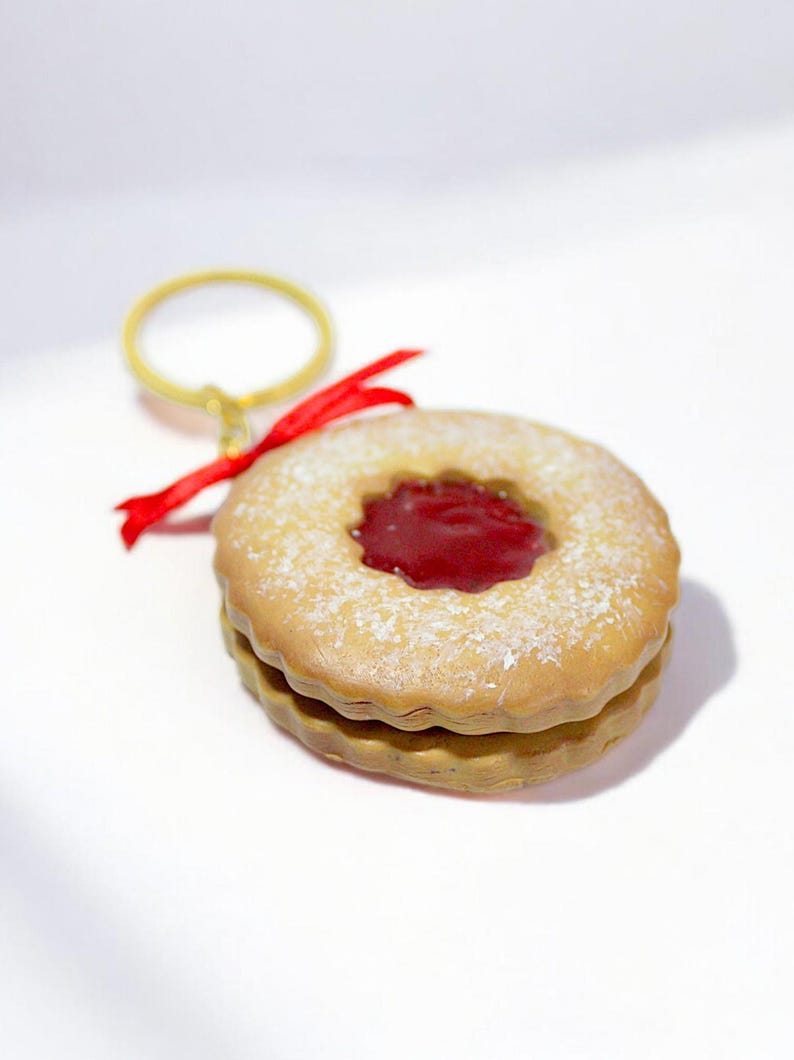 May include: A round, two-layered cookie keychain with a red jam center and a dusting of powdered sugar. The keychain has a gold ring and a red ribbon attached.