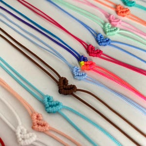 May include: A collection of colorful friendship bracelets with heart-shaped knots. The bracelets are made with different colors of string, including red, pink, blue, green, and yellow.