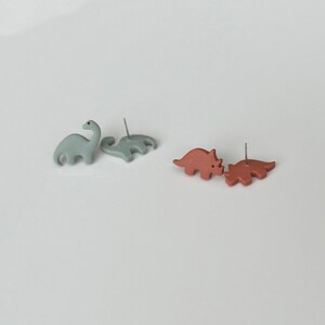 May include: Two pairs of dinosaur-shaped stud earrings. One pair is light blue, featuring a long-necked Brontosaurus and a Stegosaurus. The other pair is reddish-brown, depicting two Triceratops. The earrings are on a white surface.