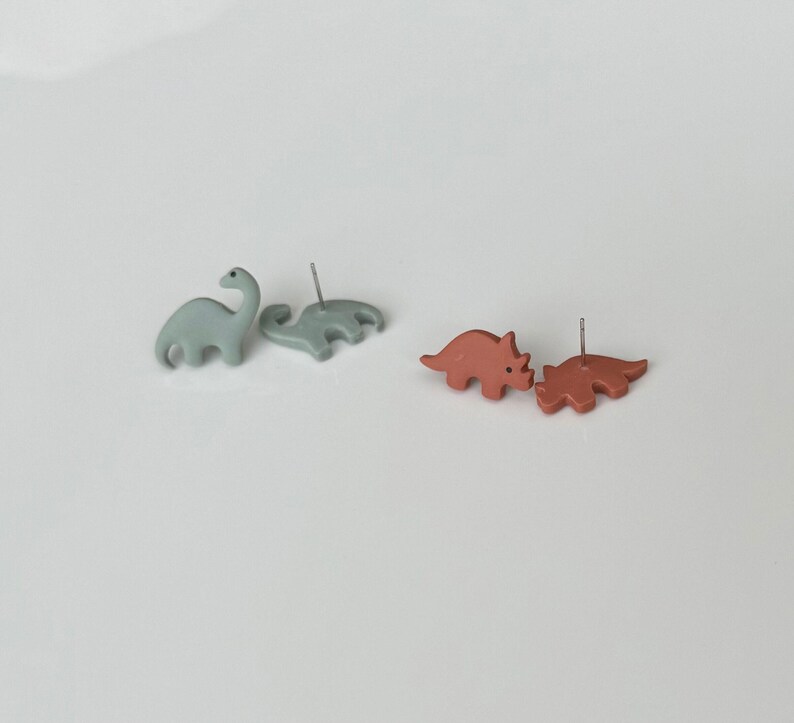 May include: Two pairs of dinosaur-shaped stud earrings. One pair is light blue, featuring a long-necked Brontosaurus and a Stegosaurus. The other pair is reddish-brown, depicting two Triceratops. The earrings are on a white surface.
