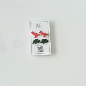 May include: A white card with four dinosaur-shaped earrings in red and green. The card has a green border and the text "Visit My Shop!" with a QR code. The earrings are small and colorful.