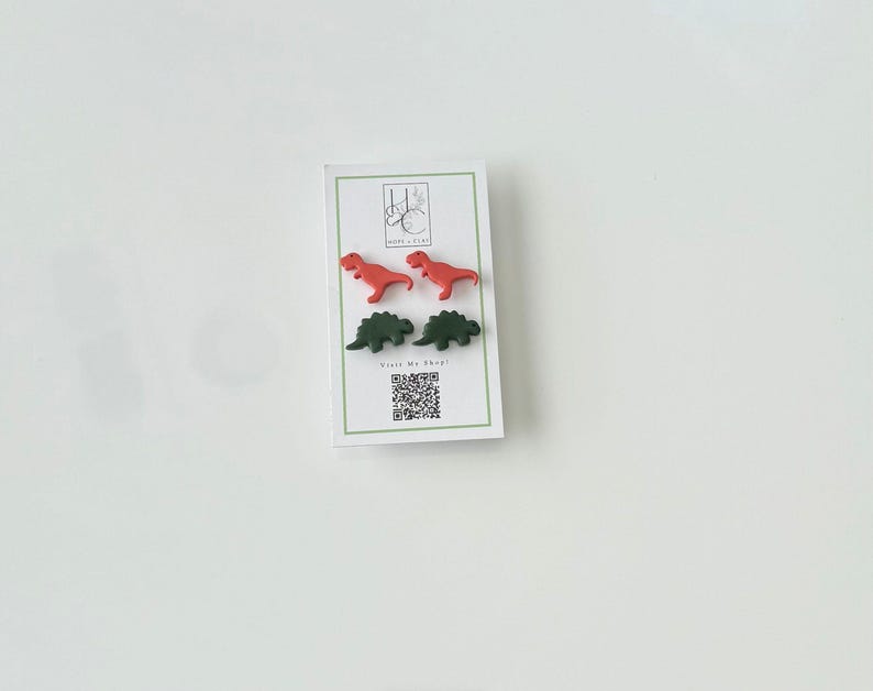 May include: A white card with four dinosaur-shaped earrings in red and green. The card has a green border and the text "Visit My Shop!" with a QR code. The earrings are small and colorful.