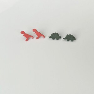 May include: Two red Tyrannosaurus Rex dinosaur-shaped earrings and two green Stegosaurus dinosaur-shaped earrings are arranged on a white surface. The earrings are small and likely made of plastic or resin.
