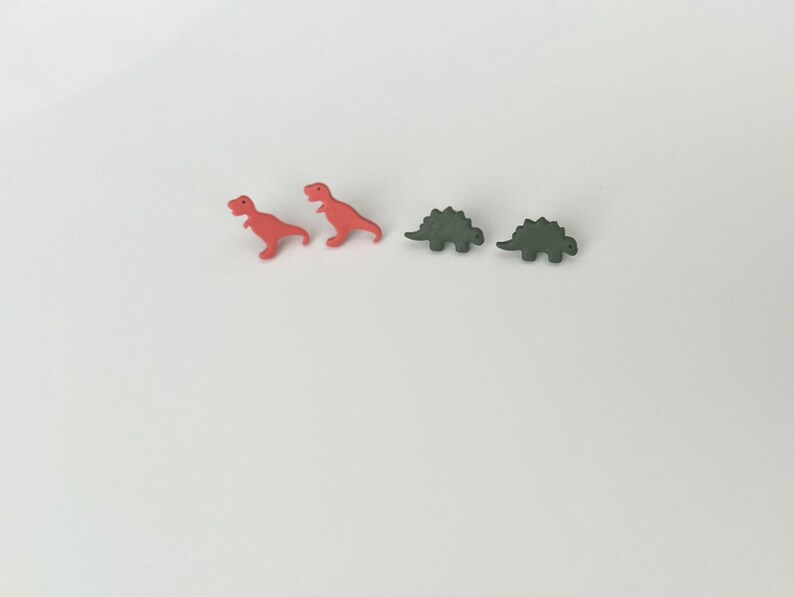 May include: Two red Tyrannosaurus Rex dinosaur-shaped earrings and two green Stegosaurus dinosaur-shaped earrings are arranged on a white surface. The earrings are small and likely made of plastic or resin.