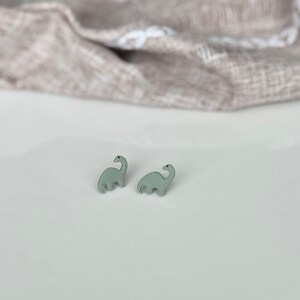 May include: A pair of light green dinosaur-shaped stud earrings. The earrings are small and feature a simple, stylized design of a Brontosaurus. The earrings are set against a white surface with a neutral-colored fabric in the background.