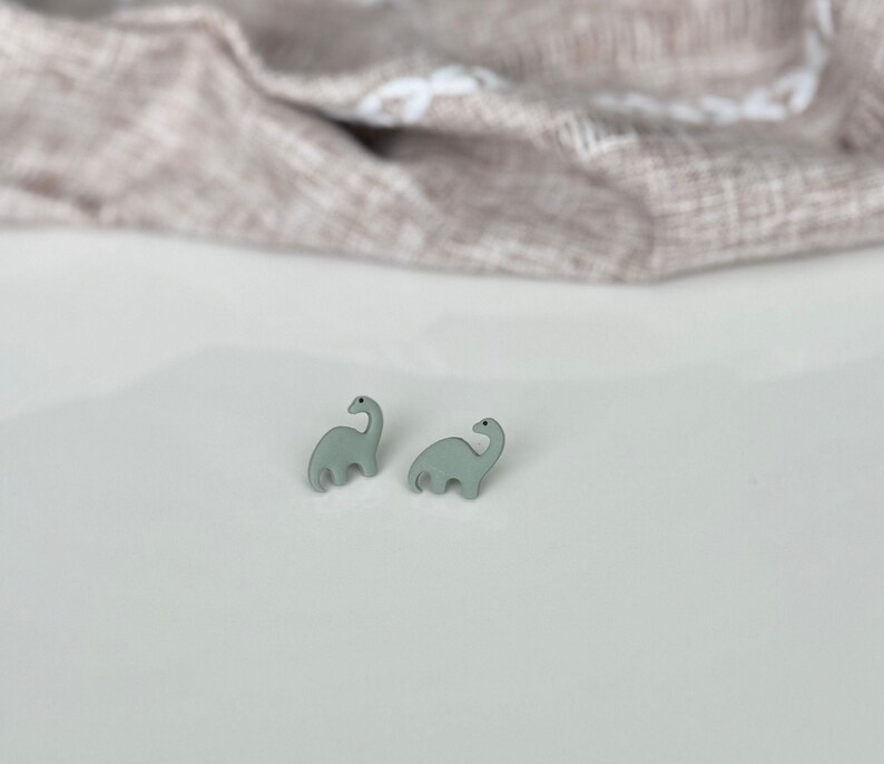 May include: A pair of light green dinosaur-shaped stud earrings. The earrings are small and feature a simple, stylized design of a Brontosaurus. The earrings are set against a white surface with a neutral-colored fabric in the background.
