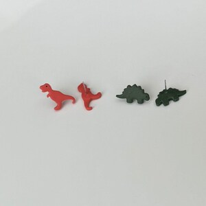 May include: A pair of red Tyrannosaurus Rex earrings and a pair of green Stegosaurus earrings. The earrings are small, with a post backing. The dinosaurs are facing different directions.