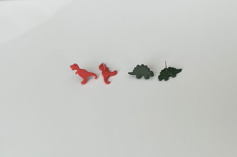 May include: A pair of red Tyrannosaurus Rex earrings and a pair of green Stegosaurus earrings. The earrings are small, with a post backing. The dinosaurs are facing different directions.