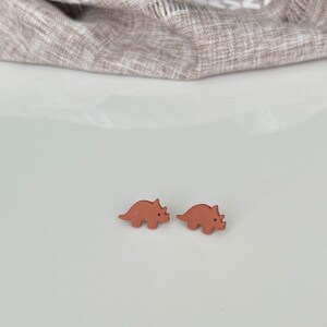 May include: Two small, terracotta-colored dinosaur stud earrings. The earrings are in the shape of a triceratops. The background is a white surface with a tan fabric in the upper portion of the image.