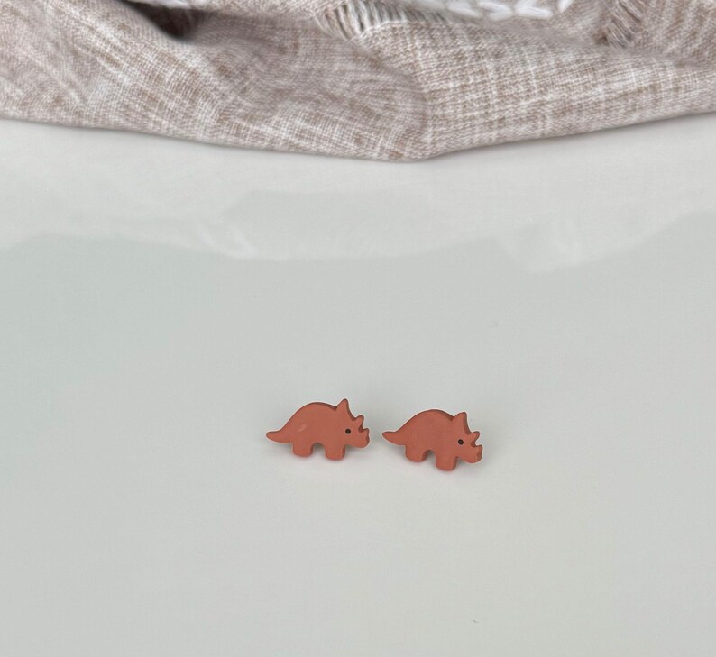 May include: Two small, terracotta-colored dinosaur stud earrings. The earrings are in the shape of a triceratops. The background is a white surface with a tan fabric in the upper portion of the image.