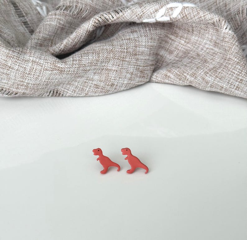 May include: Two small, red Tyrannosaurus Rex dinosaur-shaped earrings. The earrings are on a white surface, with a tan and white patterned fabric in the background.