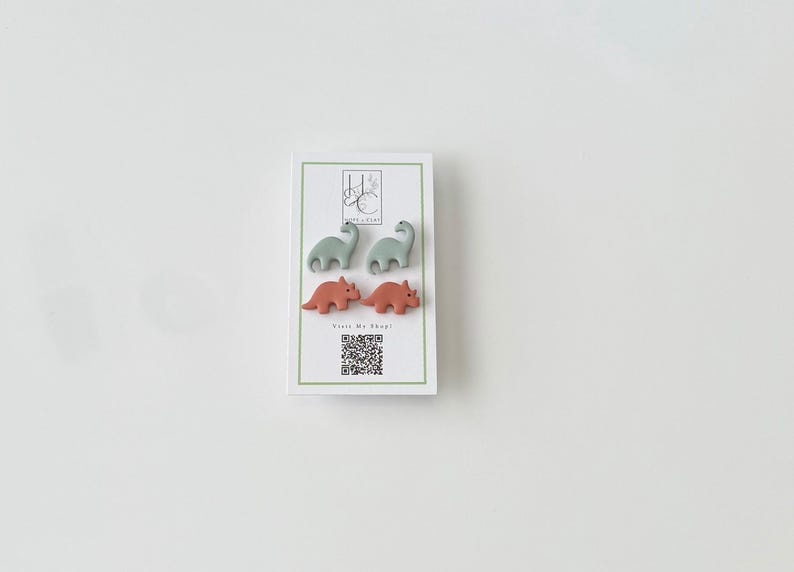 May include: A white card with two pairs of dinosaur-shaped accessories. Two green Brontosaurus and two orange Triceratops are displayed. The card has a logo and the text "Visit My Shop!" with a QR code.