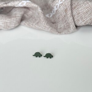 May include: Two dark green dinosaur-shaped earrings. The earrings are small, with a textured appearance, and are set against a white surface. A tan fabric with a subtle pattern is draped in the background.