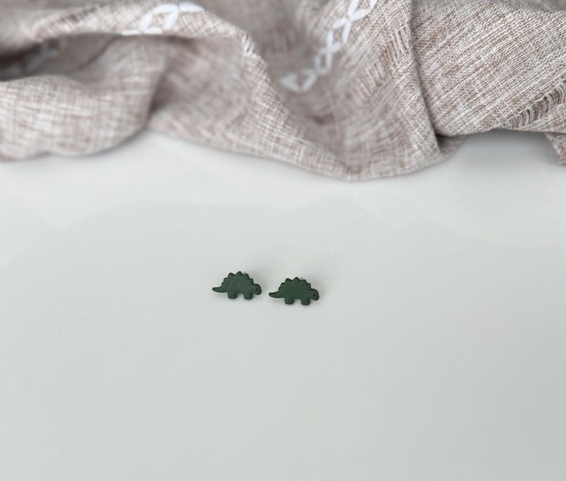 May include: Two dark green dinosaur-shaped earrings. The earrings are small, with a textured appearance, and are set against a white surface. A tan fabric with a subtle pattern is draped in the background.