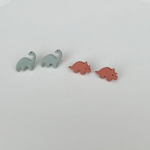 May include: Four small dinosaur-shaped objects. Two are light blue Brontosaurus figures, and two are coral-colored Triceratops figures. The dinosaurs are arranged in a group on a white surface.