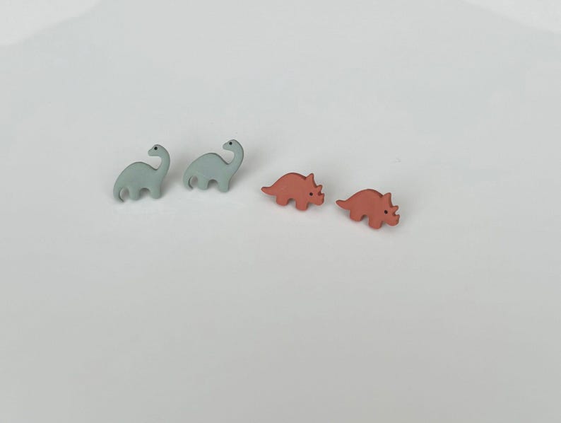 May include: Four small dinosaur-shaped objects. Two are light blue Brontosaurus figures, and two are coral-colored Triceratops figures. The dinosaurs are arranged in a group on a white surface.