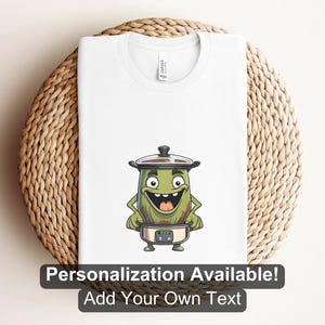 May include: White t-shirt featuring a cartoon character designed as a green slow cooker with a happy face. The shirt has the text "Personalization Available! Add Your Own Text" at the bottom. The shirt is displayed on a woven, circular surface.