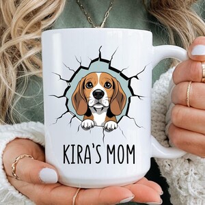 May include: A white ceramic mug featuring a graphic illustration of a beagle dog peeking through a cracked wall effect. Below the dog, the text KIRA'S MOM is printed in a simple black sans-serif font. The mug has a standard handle and smooth finish.