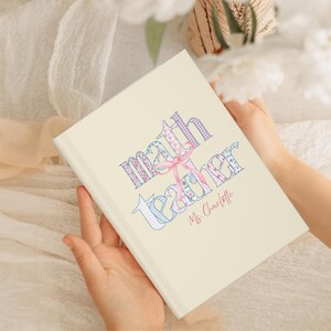 May include: A cream-coloured notebook featuring the words "Math Teacher" in a patchwork design, embellished with a pink bow. The name "Ms. Charlotte" is written below in a delicate script.