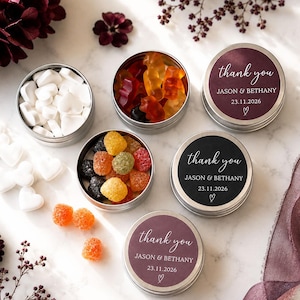 May include: Three round metal tins filled with colourful gummy sweets and white heart-shaped sweets. Two tins are closed with labels reading thank you, JASON & BETHANY, 23.11.2026, and a heart icon. One tin has a black label, two have dark purple labels.
