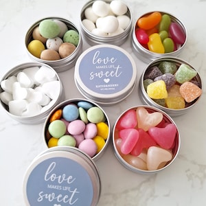 May include: A collection of small, round, silver tins filled with various candies. Some tins contain white heart-shaped candies, while others hold colorful jelly beans, pastel-colored chocolates, and sugared almonds. The lids of some tins feature the text "love makes life sweet".