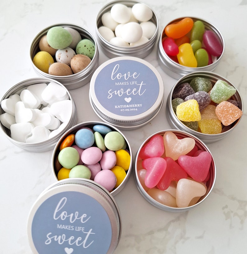 May include: A collection of small, round, silver tins filled with various candies. Some tins contain white heart-shaped candies, while others hold colorful jelly beans, pastel-colored chocolates, and sugared almonds. The lids of some tins feature the text "love makes life sweet".